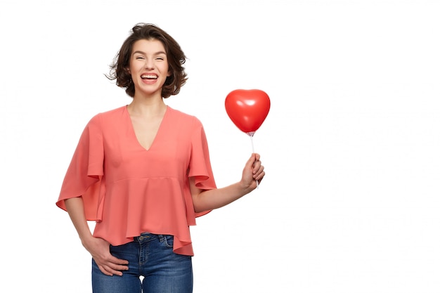 Eine schöne Brunettefrau mit einem breiten Lächeln in den Jeans und in einer rosa Jacke, die rote Ballone in Form eines Herzens hält