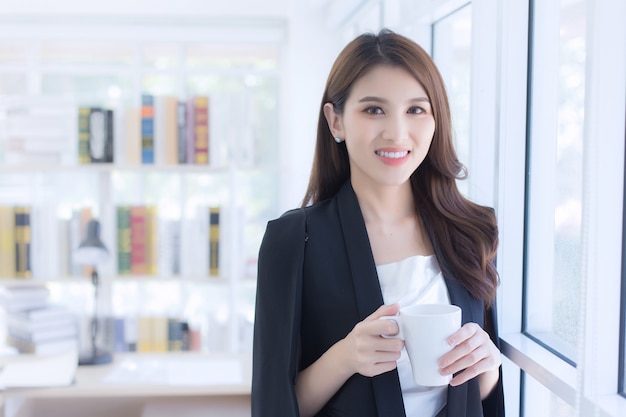 Eine schöne Bürodame, die eine Kaffeetasse am Fenster hält und glücklich am Arbeitsplatz steht