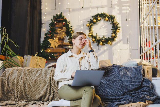 Eine schöne Frau arbeitet zu Hause in den Neujahrsferien. Tragen Sie bequeme Hauskleidung. Schauen Sie sich einen Laptop an. Arbeiten Sie aus der Ferne für das neue Jahr