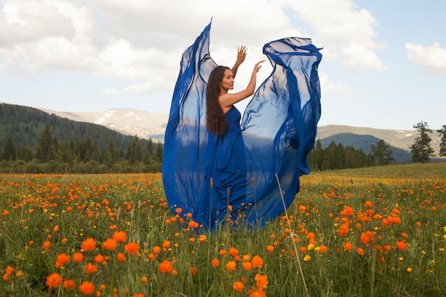 Eine schöne Frau asiatischen Aussehens in einem blauen Kleid spaziert im Sommer auf einer grünen Wiese mit orangefarbenen Blumen im Altai-Gebirge. Konzept Schönheit, Tourismus.