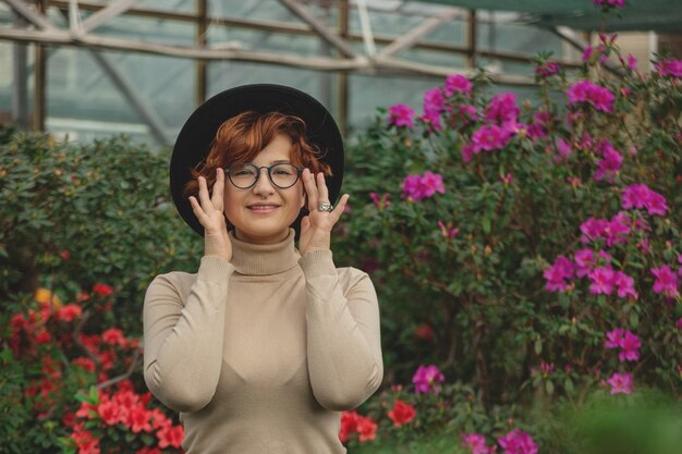 Eine schöne Frau in Brille und Hut lächelnd zwischen den grünen Pflanzen und Blumen.