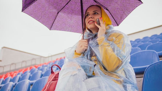 Eine schöne Frau in einem Hoodie und einem Regenmantel aus Plastik, die während des Regens mit einem Handy auf dem Podium sitzt und einen Regenschirm in der Hand hält