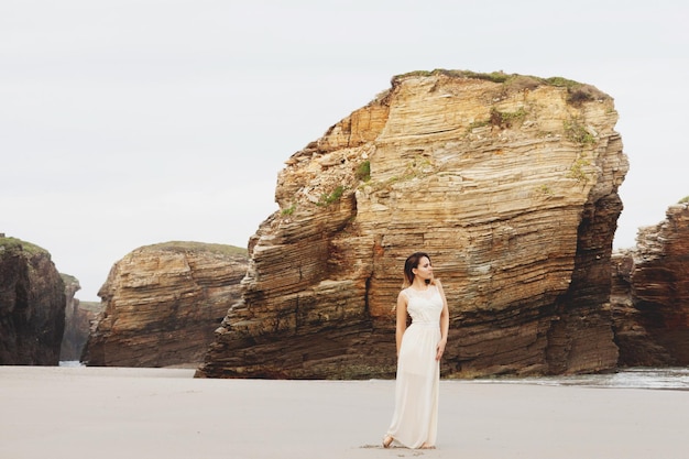 Eine schöne Frau in einem langen romantischen Kleid geht barfuß an einem wunderschönen Strand mit hohen Klippen spazieren