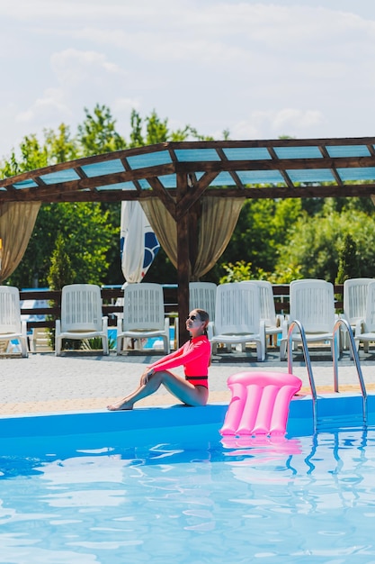 Eine schöne Frau in einem rosafarbenen Badeanzug und mit einer aufblasbaren Matratze steht in der Nähe eines großen Pools Sommerferien in einem Hotel mit Swimmingpool Junges Mädchen in Badeanzug und Sonnenbrille