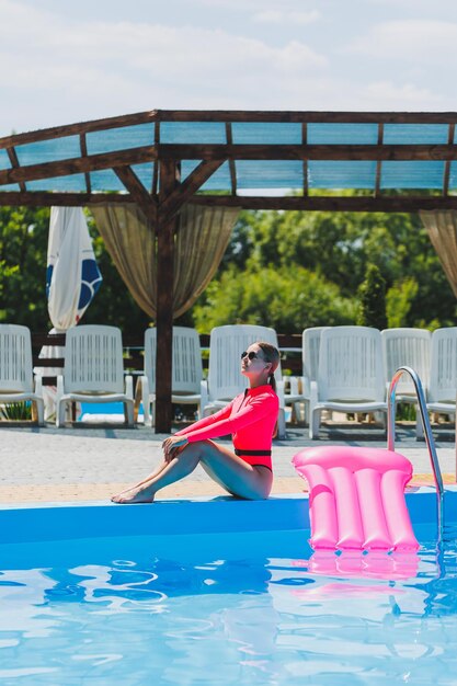 Eine schöne Frau in einem rosafarbenen Badeanzug und mit einer aufblasbaren Matratze steht in der Nähe eines großen Pools Sommerferien in einem Hotel mit Swimmingpool Junges Mädchen in Badeanzug und Sonnenbrille