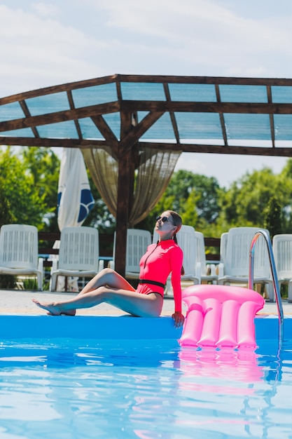 Eine schöne Frau in einem rosafarbenen Badeanzug und mit einer aufblasbaren Matratze steht in der Nähe eines großen Pools Sommerferien in einem Hotel mit Swimmingpool Junges Mädchen in Badeanzug und Sonnenbrille