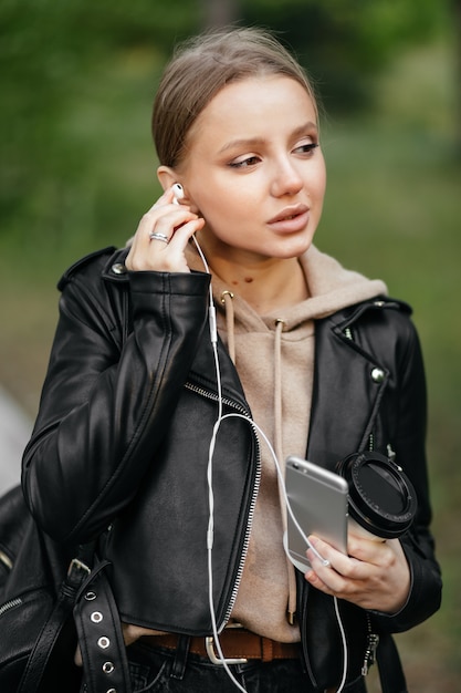 Eine schöne Frau in einer Lederjacke geht durch den Park und steckt Kopfhörer in ihre Ohren, um Musik vom Telefon zu hören