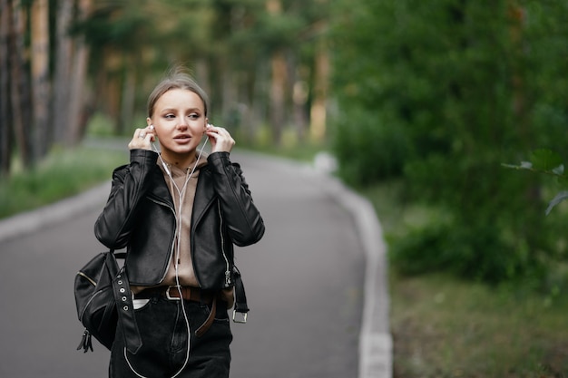 Eine schöne Frau in einer Lederjacke geht durch den Park und steckt Kopfhörer in ihre Ohren, um Musik vom Telefon zu hören