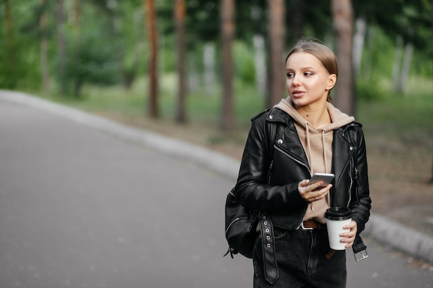 Eine schöne Frau in einer Lederjacke geht in den Park und lächelt hübsch, während sie mit einem Telefon und Kaffee in den Händen zur Seite schaut
