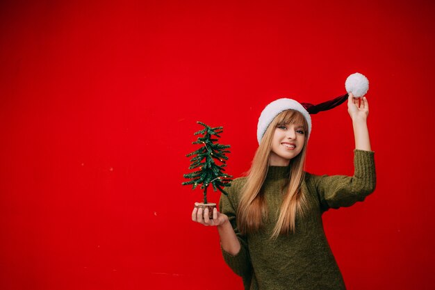 Eine schöne Frau in einer Weihnachtsmütze hält einen kleinen Weihnachtsbaum in ihren Händen auf rotem Hintergrund