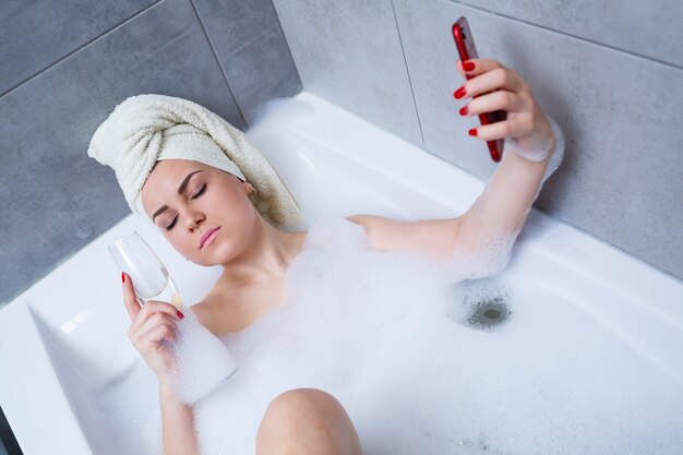 Eine schöne Frau mit einem Handtuch auf dem Kopf liegt mit einem Glas Champagner in einem weißen Bad und macht ein Selfie am Telefon. Entspannen Sie sich nach einem anstrengenden Tag. Entspannende Spa-Behandlung