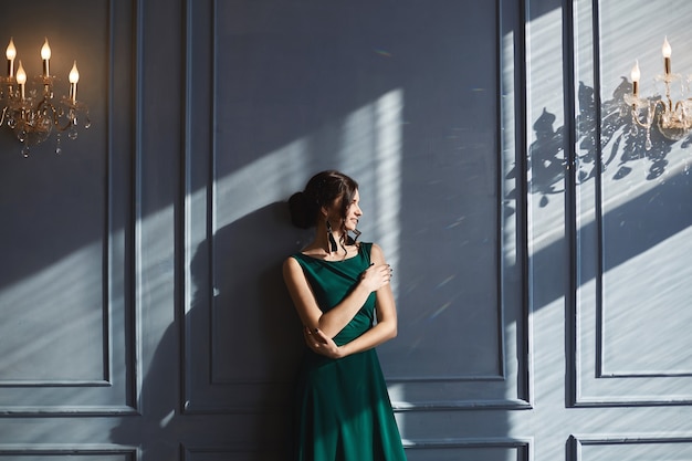 Eine schöne Frau mit eleganter Frisur im grünen Abendkleid steht an der blauen Wand mit Sonnenlicht und Schatten
