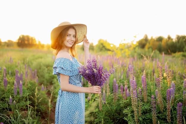 Eine schöne Frau mit Hut und Kleid hält einen Strauß Lupinen und lächelt