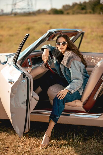 Eine schöne Frau sitzt auf einem Cabrio auf einem Feld mit Sonnenbrille, gekleidet in Jeans und einer Pelzjacke, schön und stilvoll.