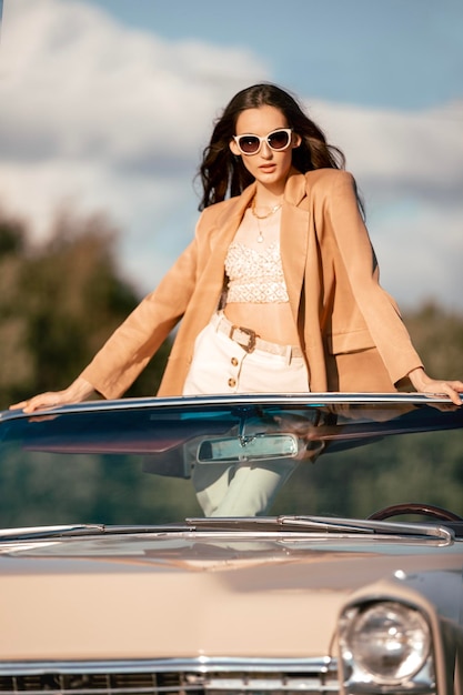 Eine schöne Frau sitzt in einem Cabriolet auf einem Feld, gekleidet in einer beigen Jacke und weißen Hose und schwarzen Sonnenbrille, schön und stilvoll.