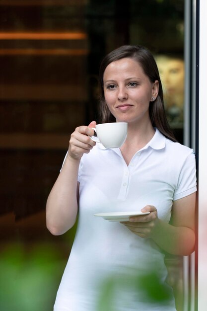 Eine schöne Frau trinkt Kaffee oder Tee aus einer weißen Tasse, die in der Tür steht, und erwartet Freunde oder...