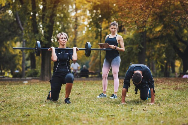 Eine schöne Frau und ihr Partner trainieren mit einem Personal Trainer im Park, gekleidet in einen schwarzen Anzug mit einem elektronischen EMS-Simulator, um ihre Muskeln zu stimulieren.