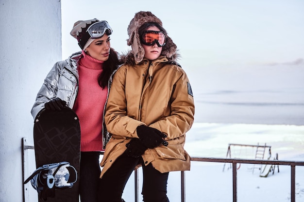 Eine schöne Frau und ihr Sohn in stilvollen warmen Kleidern, die mit einem Snowboard neben einem Geländer am schneebedeckten Strand stehen