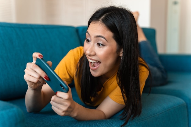 eine schöne fröhliche emotionale erfreute brünette junge frau auf dem sofa drinnen zu hause spielen spiele per handy.