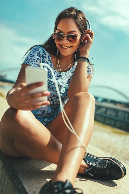 Eine schöne, fröhliche junge Frau hört die Musik vom Smartphone und genießt an einem schönen Sommertag am Ufer des Stadtflusses.