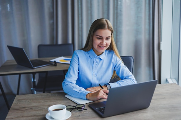 Eine schöne Geschäftsfrau in einem Hemd und einer Brille sitzt mit einem Laptop im Büro am Tisch Managerin mit Brille am Arbeitsplatz im modernen Büro Arbeitstag am Computer