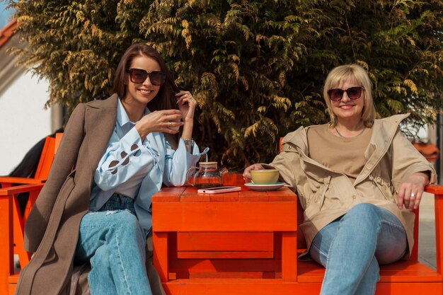 Eine schöne, glückliche ältere Mutter mit einem Lächeln und eine fröhliche, hübsche Tochter mit Sonnenbrille in modischer Kleidung sitzen draußen in einem Café und trinken Kaffee und Tee an einem sonnigen Frühlingstag