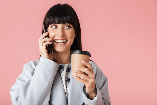 Eine schöne glückliche Frau, die isoliert über hellrosa Wand posiert und mit dem Handy spricht, das Kaffee hält.