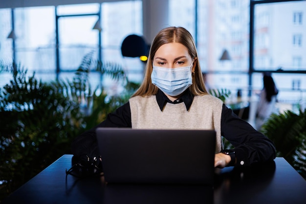 Eine schöne, intelligente Geschäftsfrau in einem großen Unternehmen hält ein Online-Meeting mit Geschäftspartnern in einer Schutzmaske während des Coronavirus sitzt sie mit einem Laptop an einem Tisch an einem Arbeitsplatz