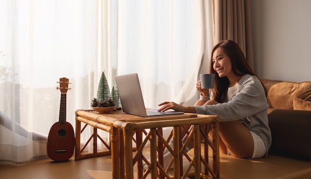 Eine schöne junge asiatische Frau, die Laptop-Computer verwendet und daran arbeitet, während sie zu Hause Kaffee trinkt