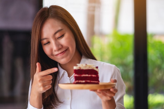 Eine schöne junge asiatische Frau, die mit dem Finger auf ein Stück roten Samtkuchen in einem Holztablett auf dem Tisch zeigt