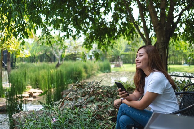 Eine schöne junge asiatische Frau mit Handy mit Laptop-Computer auf dem Tisch im Park