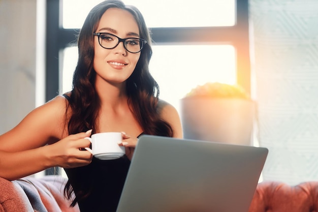 Eine schöne junge europäische Bloggerin sitzt mit einer Tasse Kaffee oder Tee an einem Laptop und lächelt