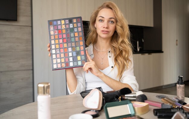 Eine schöne junge Frau bloggt über Make-up und Schönheitsprodukte. Wunderschöne Dame, die Make-up-Tutorial mit Make-up-Lidschatten-Palette abnimmt. Lächelndes Mädchen lehrt Make-up mithilfe der Webcam-Ansicht ihres Telefons