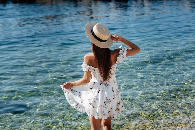 Eine schöne junge Frau in einem Hut und einem leichten Kleid mit dem Rücken geht an einem sonnigen Tag am Meer entlang vor dem Hintergrund riesiger Felsen Tourismus- und Urlaubsreisen