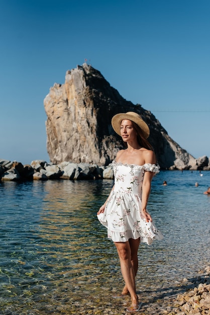 Eine schöne junge Frau in einer Hutbrille und einem leichten Kleid geht an einem sonnigen Tag vor dem Hintergrund riesiger Felsen am Meeresufer entlang Tourismus- und Touristenreisen