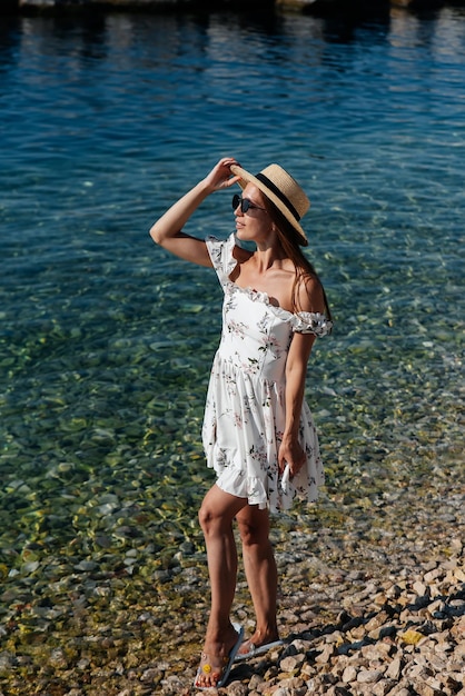 Eine schöne junge Frau in einer Hutbrille und einem leichten Kleid geht an einem sonnigen Tag vor dem Hintergrund riesiger Felsen am Meeresufer entlang Tourismus- und Touristenreisen