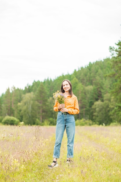Eine schöne junge Frau in einer orangefarbenen Bluse, Jeans mit Blumen an den Händen steht und lächelt in die Kamera auf dem Feld vor dem Hintergrund eines grünen Waldes