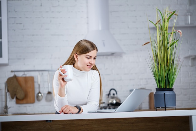 Eine schöne junge Frau mit blonden Haaren in einem weißen Rollkragenpullover steht in einer weißen Küche und arbeitet an einem Laptop, sie trinkt Kaffee. Arbeiten Sie aus der Ferne von zu Hause aus