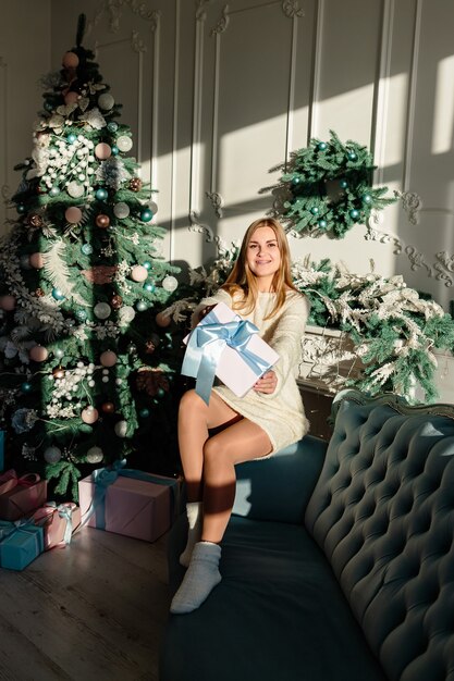 Eine schöne junge Frau mit blonden Haaren in heller Kleidung sitzt auf dem Sofa und hält ein verpacktes Geschenk mit einer Schleife in den Händen. Zeit, Weihnachtsgeschenke zu öffnen. Bald ist Silvester