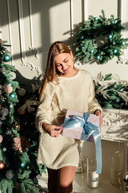 Eine schöne junge Frau mit blonden Haaren steht neben einem Weihnachtsbaum mit einem Geschenk in den Händen. Bald kommt das neue Jahr. Weihnachtsstimmung in einem gemütlichen Zuhause