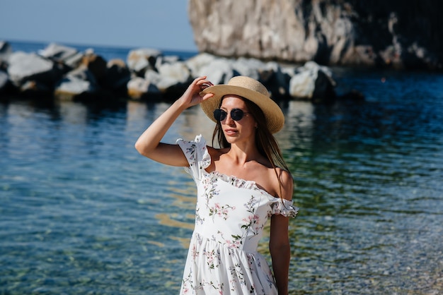 Eine schöne junge Frau mit Hut, Brille und einem leichten Kleid geht an einem sonnigen Tag vor dem Hintergrund riesiger Felsen am Meer entlang. Tourismus und touristische Reisen.