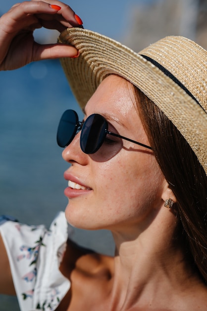 Eine schöne junge Frau mit Hut, Brille und einem leichten Kleid geht an einem sonnigen Tag vor dem Hintergrund riesiger Felsen am Meer entlang. Tourismus und touristische Reisen.