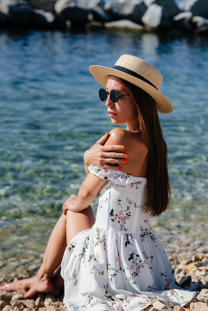 Eine schöne junge Frau mit Hut, Brille und einem leichten Kleid sitzt an einem sonnigen Tag am Meeresufer vor dem Hintergrund riesiger Felsen. Tourismus und touristische Reisen.