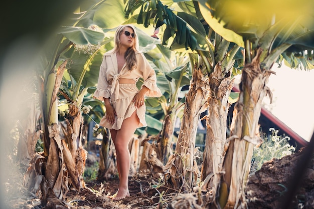 Eine schöne junge Frau mit langen blonden Haaren europäischer Erscheinung steht in der Nähe der Bananenbäume Mädchen im tropischen Wald an einem sonnigen Sommertag