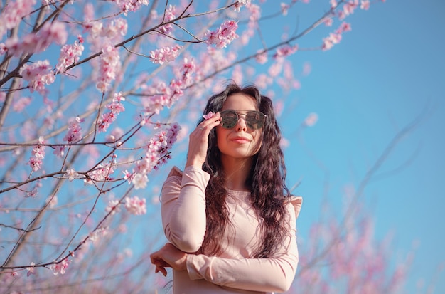 Eine schöne junge frau mit verspiegelter sonnenbrille nahe einem blühenden frühlingskirschblütenbaum. die idee und das konzept von erneuerung, selbstfürsorge, gesundheit und glück