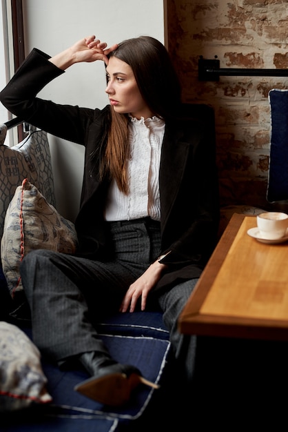 Eine schöne junge Frau sitzt in einem Café hinter einem Glas mit einer Tasse Kaffee und schaut nachdenklich aus dem Fenster.