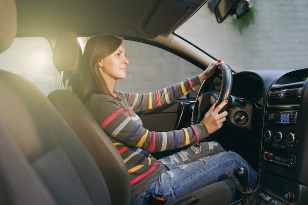 Foto eine schöne junge, glücklich lächelnde europäische braunhaarige frau mit gesunder, sauberer haut in einem gestreiften t-shirt sitzt in ihrem auto mit schwarzem interieur. reise- und fahrkonzept.