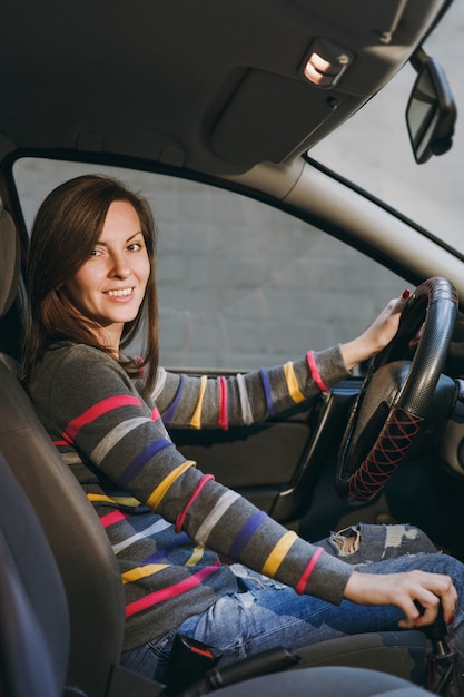 Eine schöne junge, glücklich lächelnde europäische braunhaarige Frau mit gesunder, sauberer Haut in einem gestreiften T-Shirt sitzt in ihrem Auto mit schwarzem Interieur. Reise- und Fahrkonzept.