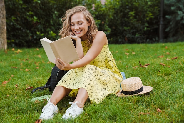 eine schöne junge hübsche optimistische frau sitzt draußen auf gras im naturpark und liest buch.