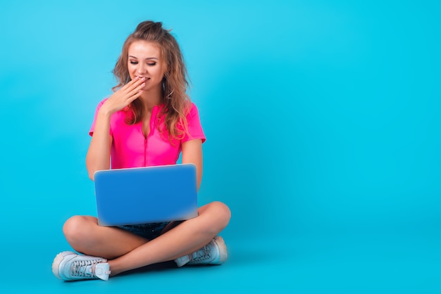 Eine schöne junge lächelnde Frau in einem hellen Outfit, die isoliert auf einem hellblauen Hintergrund sitzt und an ihrem Laptop arbeitet. Emotionales Gesicht. Horizontale Studioaufnahme in voller Länge. Platz kopieren.